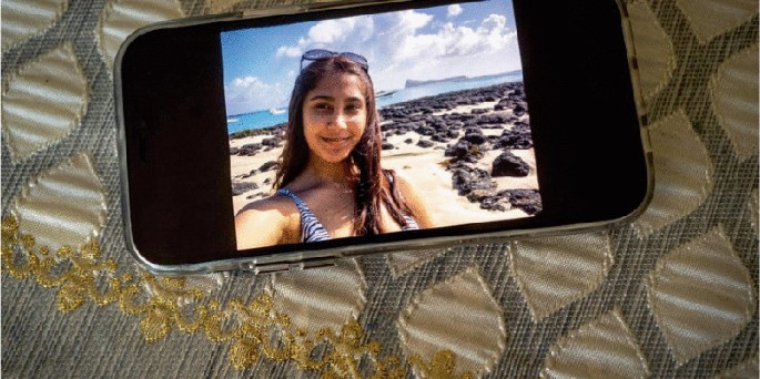 A close-up of a smartphone displays a photo of a woman on a rocky beach with the ocean and the sky in the background. The smartphone is placed on top of a textured, golden-embroidered cloth.