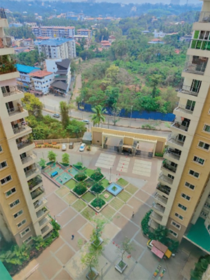 An aerial view photograph of Brigade Pinnacle with a well-defined parking area, lush gardens, and elegant fountains. In the backdrop, a vegetative forest and residential buildings.