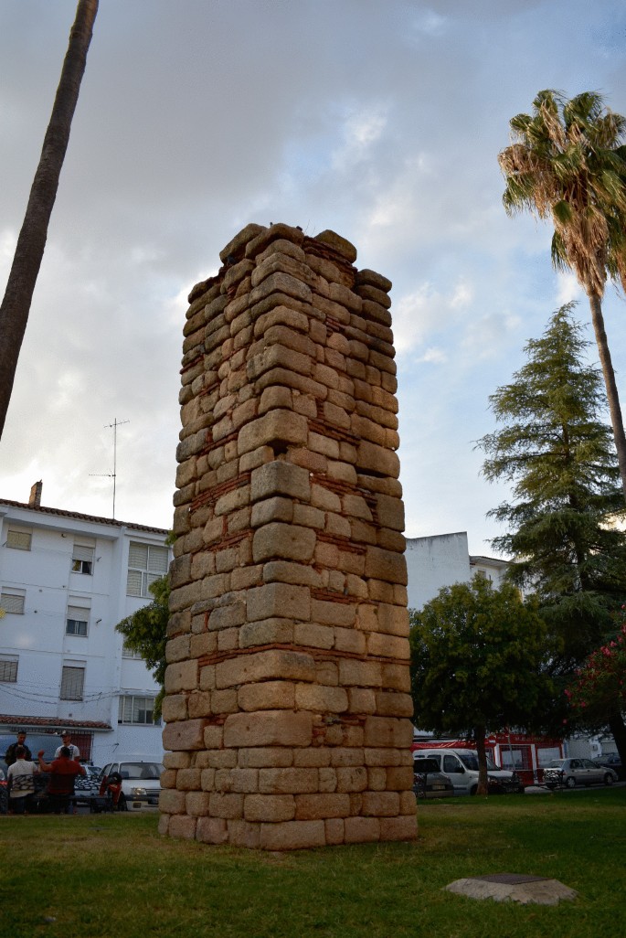 A photograph of a tall, cuboid column made of stone bricks. A few gaps have been filled with flat, clay material. The column stands on the lawn in front of a building.