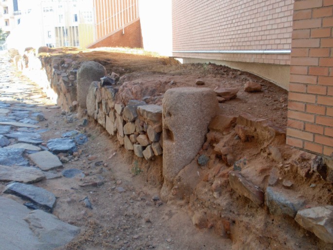 A photograph of a short platform made of mud and large stone bricks that stands on the side of modern brick buildings. The pathway near it is paved with flat rocks embedded into the ground. 2 short stone pillars with smoothened and curved edges are present.