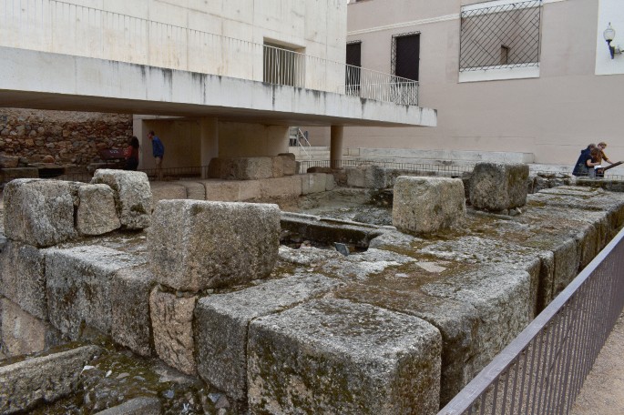 A photograph of a short structure that resembles a platform. It is made of large stone blocks and lies near a modern building. A few of the stones are covered in moss.