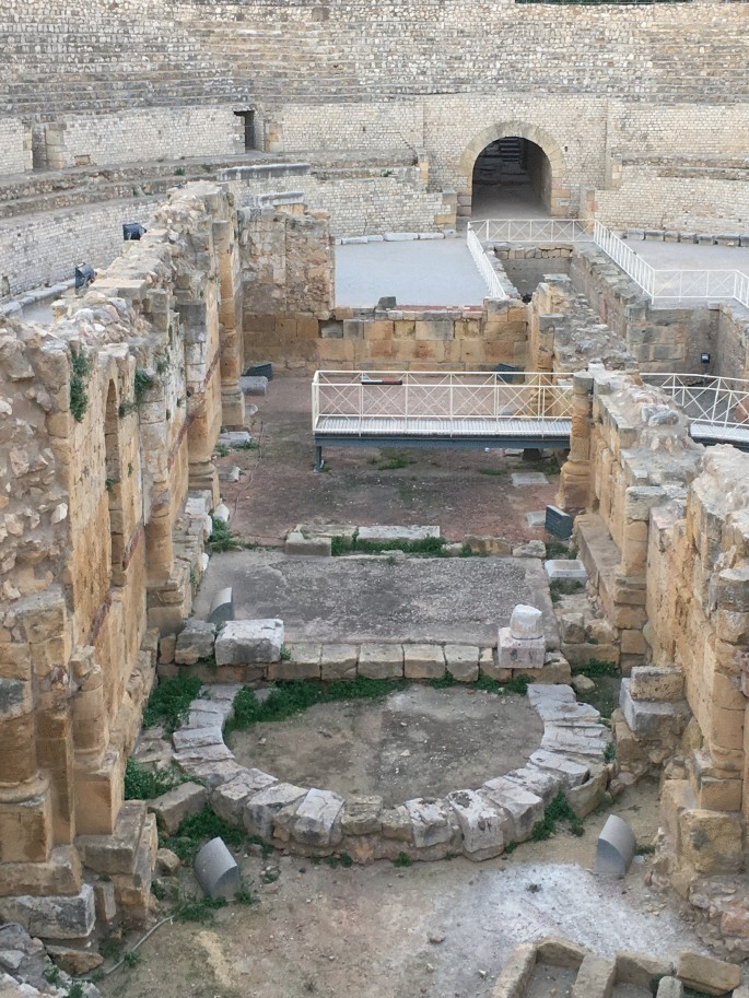 A photograph of a large amphitheater made of stone blocks. It has seats in the form of steps that surround the open area in the center. A stone parapet wall structure is present in the center. Modern railings are present around a part of the wall. An entrance is in the far end.