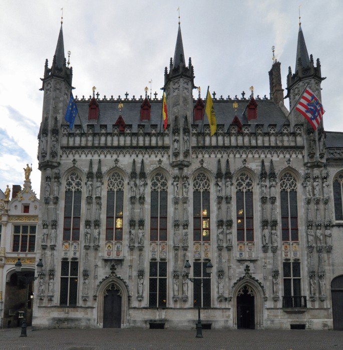 A photo of the town hall in Bruges presents a large building with many windows and a clock tower. Architectural elements include stepped gables and corner turrets.