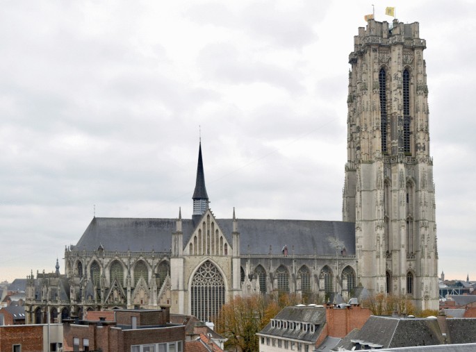A photograph of the Tower of Saint Rumbold's in Mechelen encompass a large building with a clock tower.