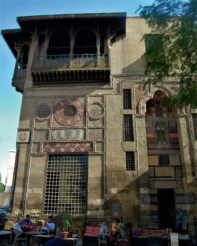 A photograph of a 2 story building for Sabil with different designs. A few people are seated in front of the building, which has a sizable grilled window at its front.