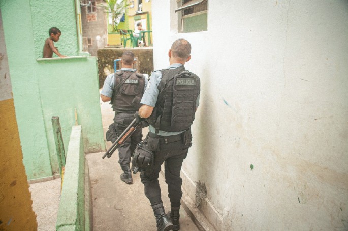 A photograph of two cops carrying firearms through a narrow pathway between two buildings. A child looks at them from one of the houses.