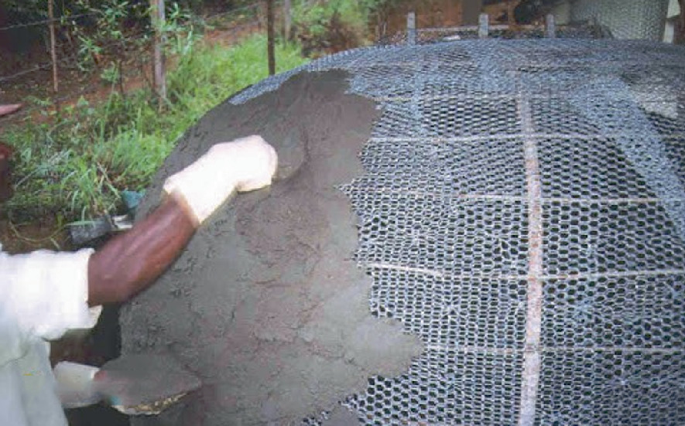 A photo of a hand applying cement onto a mesh structure dome, showcasing the construction process in a visually engaging manner.