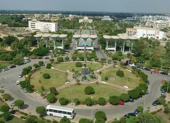 An aerial view of a well-maintained main entrance of the Suez Canal University, surrounded by roads and buildings. It features a circular layout with manicured lawns, pathways, and a prominent monument at its center. The main gate has 2 entry roads with tall structures.
