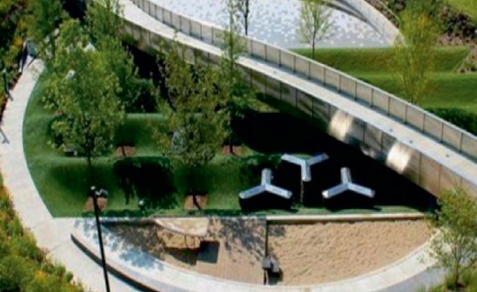 An aerial view of a modern, landscaped outdoor area. It features a curved concrete walkway, bordered by manicured grass and trees, and surrounds a lower section featuring unique benches arranged on a sandy surface. The other side of the walk has a small pond surrounded by manicured grass.