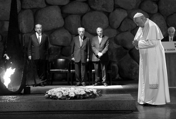 A photo of Pope Francis in side view standing with folded hands, a bowed head, in front of a tomb with a wreath on top. A flame burns to the left and 3 men in suits stand at a distance in the background watching the Pope.