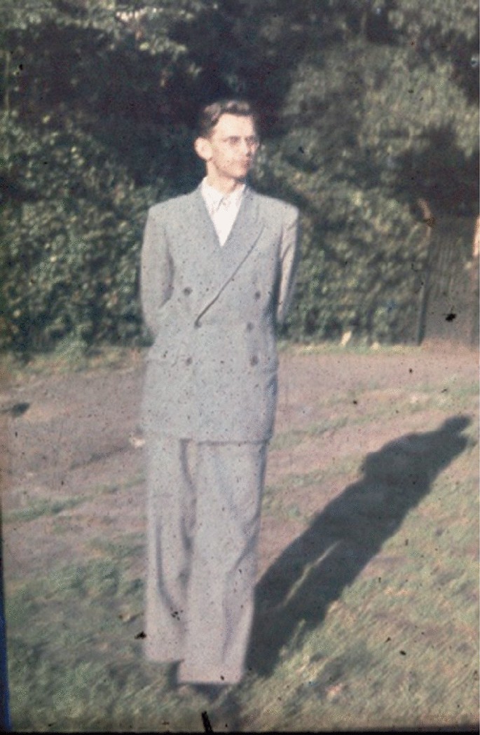 A photo of a person who stands outdoors, wearing a coat and glasses, with hands clasped behind their back. He gazes toward the left side, with trees and bushes in the background.