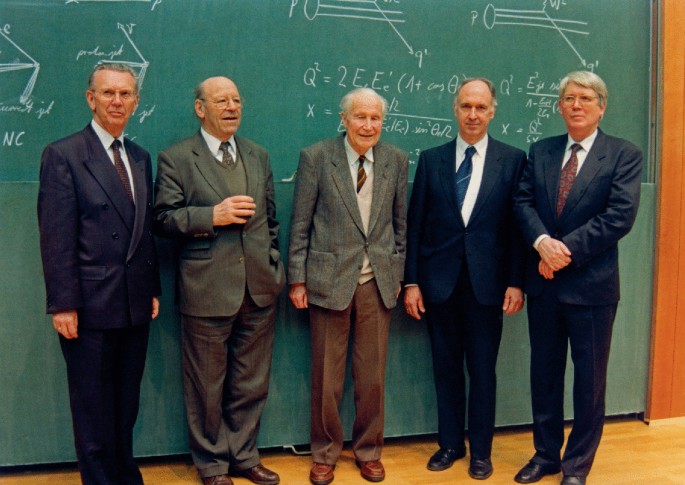 A photograph of chairs of the D E S Y directorate, Herwig Schopper, Wolfgang Paul, Willilbald Jentschke, and Volker Soergel. They stand in front of a green board for the picture.