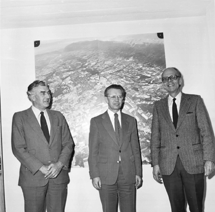 A photograph of Herwig, John Adams, and Leon van Hove. They stand in front of a large aerial photograph on the wall.
