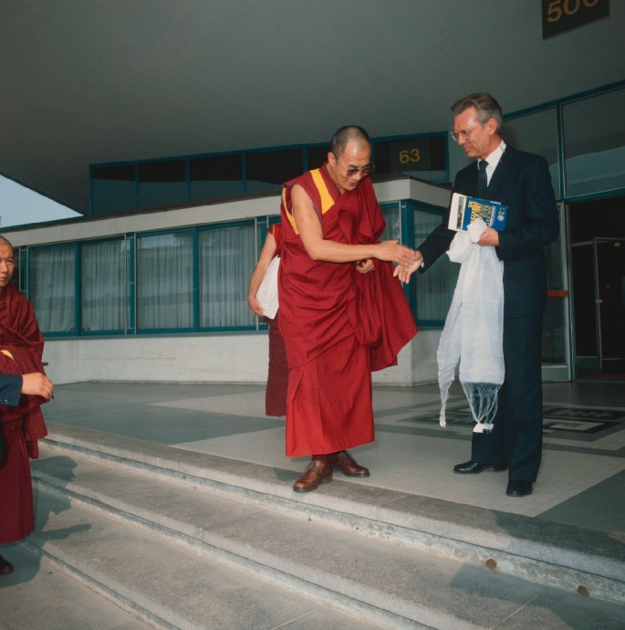 A photograph of Herwig welcoming the monk Dalai Lama at the entrance of a C E R N. Dalai Lama is wearing a Buddhist monk's attire, while Herwig is wearing a suit.
