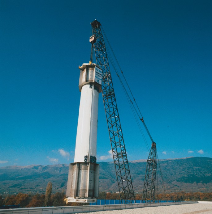 A photograph presents a construction crane lifting the support tube. There is a mountain range in the background.