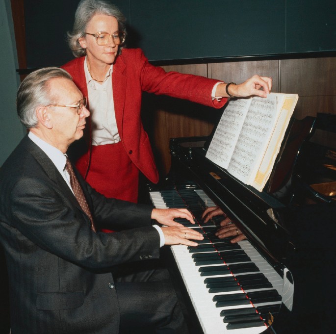 A photograph of a man and a woman. The man is playing the piano and the woman is looking at the musical notes in front of him.