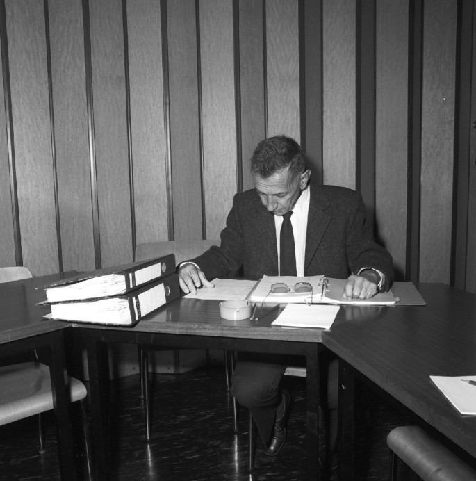 A monochrome photograph of Giuseppe Cocconi. He is sitting at a table and reading a book. There are some document files on the table.
