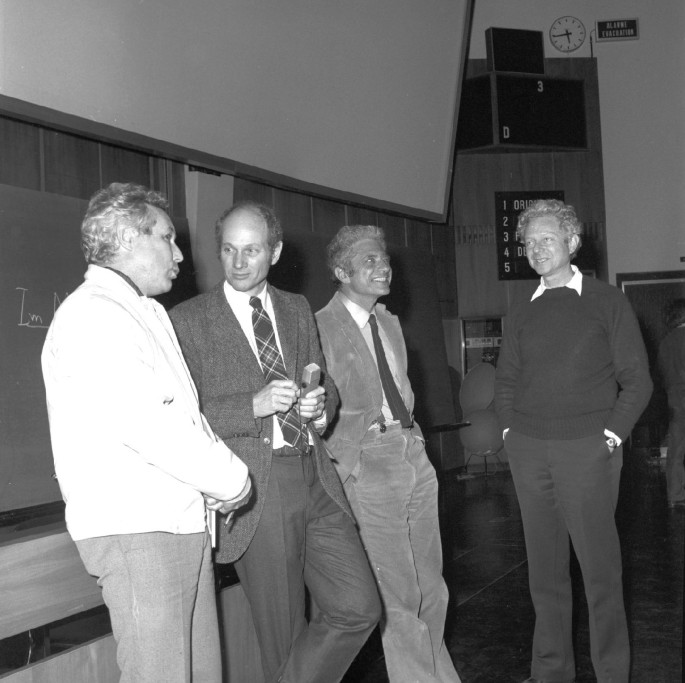 A photograph of four people standing in a hall room. They are talking and smiling with each other.