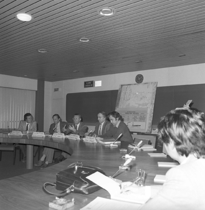 A photograph of a group of people sitting at a round table in a hall room. There are nameplates, microphones and documents on the table.
