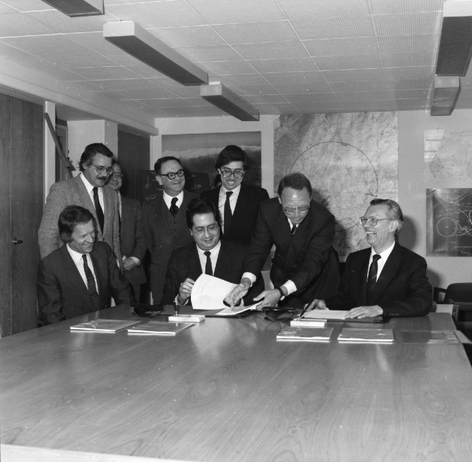 A photograph displays the Portuguese minister sitting at the table along with other people in a hall room. The minister is looking at a document while having a conversation with others. There are some documents on the table.