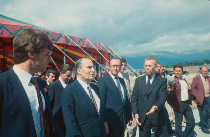 A photograph of Herwig and the president of France along with other people discussing the L E P in an open area. There is a tent in the background.