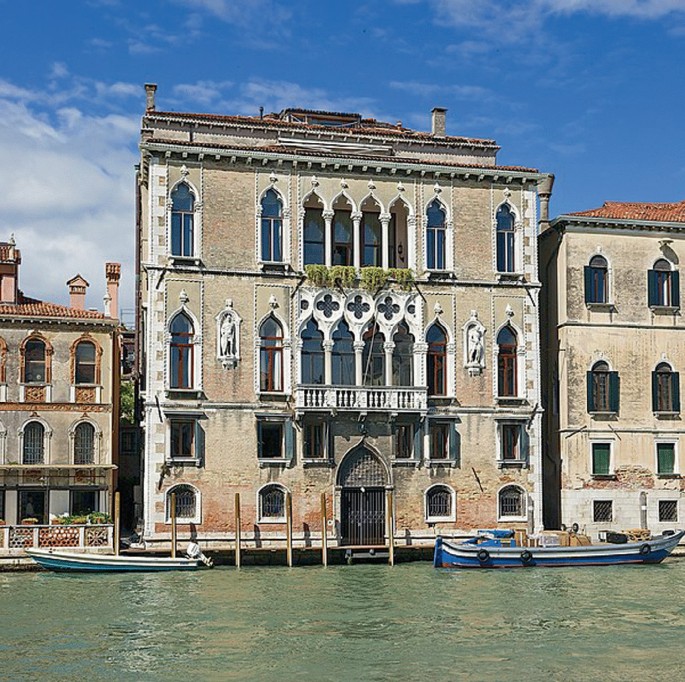 A photo of a 3-storied building with vintage architecture. It has a canal frontage with a few boats anchored. The building has large balconies and long verandas with arched hallways.