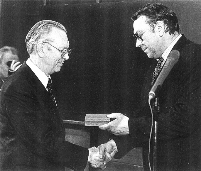 A photo presents the side-view of 2 men. The one on the right hands over a small rectangular box with his left hand to the man on the left while shaking his right hand with the latter.