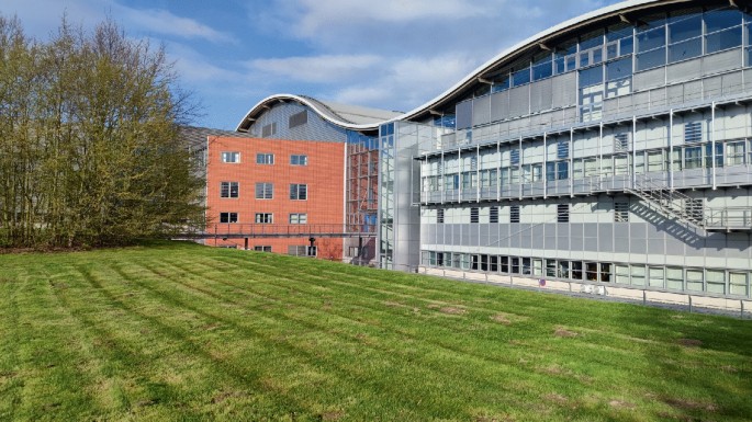 A photo presents the long shot of a large and wide multi-storied building with a roof that resembles a sine wave. It has a wide and well-mowed lawn frontage.