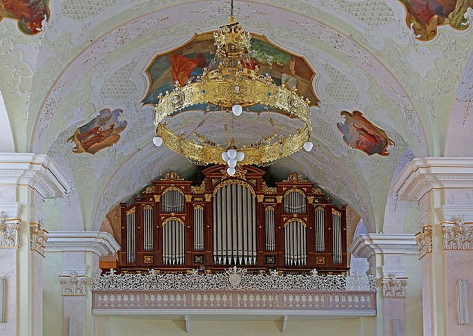 A photo of a large pipe organ installed at a height close to the ornate ceiling of a monastery. The roof curves into a dome with a chandelier hanging from its center.