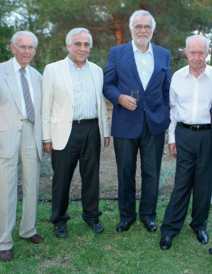 A photo of 4 men in suits stand posing for the camera in an open garden space.