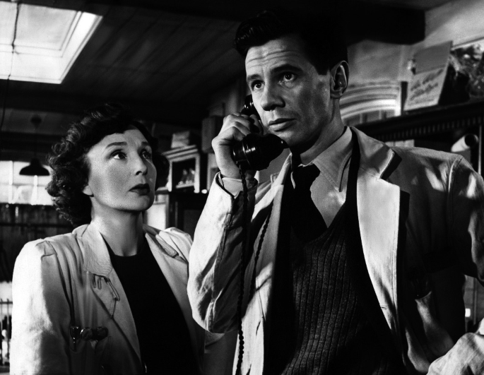 James Donald holds a telephone receiver to his ear while Googie Withers stands next to him and looks at him, both dressed as doctors. The scene is set in a hospital.