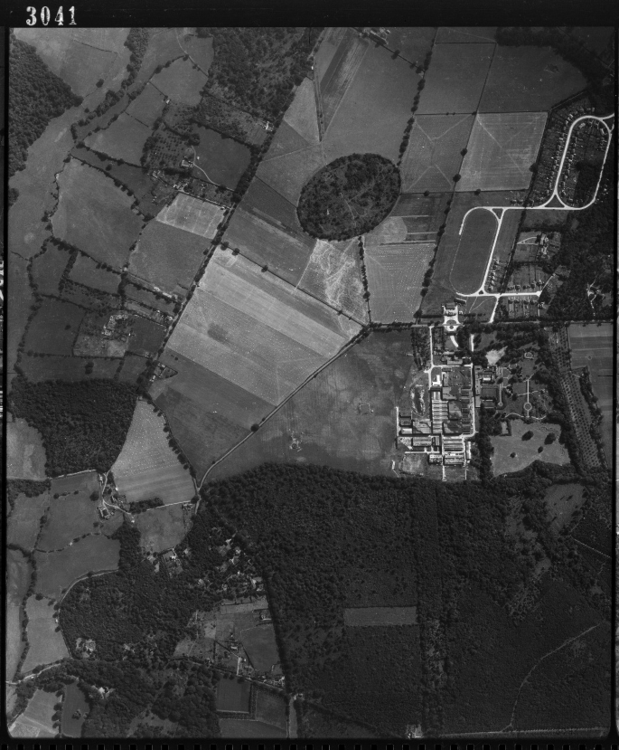 A grayscale aerial view shot of the Pinewood Studios from 1945. It consists of a patchwork of agricultural fields, forested areas, and a built structure with adjacent roads. The fields vary in shape and shade. A prominent dark circular forested area is at the top center.