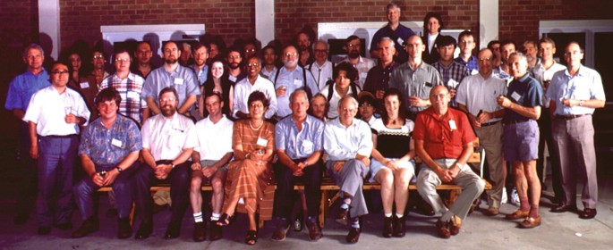 A photo of a group of men and women, a few sitting in front and others standing behind against a brick wall.