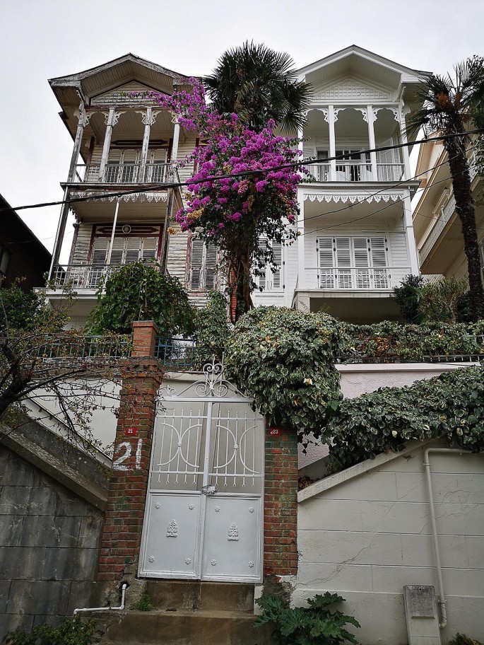 A close-up photograph of a 2-tier mansion with an inverted V-shaped roof. Various trees and flowering plants are inside the mansion and at the entrance. The entrance gate has brick pillars numbered 21 and 23.