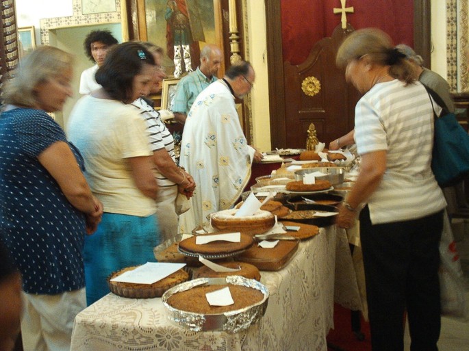 A photograph of 8 people who gaze at a long vertical table loaded with baked cakes in the church. A paper slip is placed over each cake.