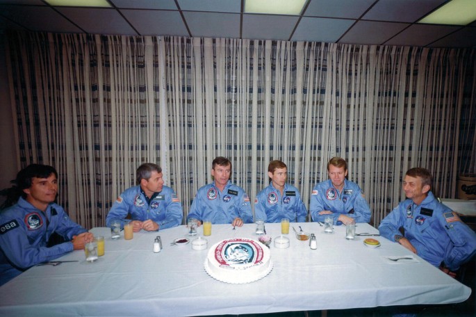 A photograph of Ulf Merbold, Bob Parker, John Young, Brewster Shaw, Byron Lichtenberg, and Owen Garriott, sitting together for breakfast.