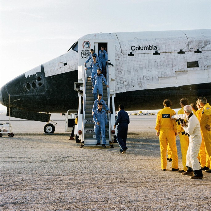 A photograph displays crewmembers coming out of the Columbia space shuttle, through the stairs. Some people are there to welcome them.