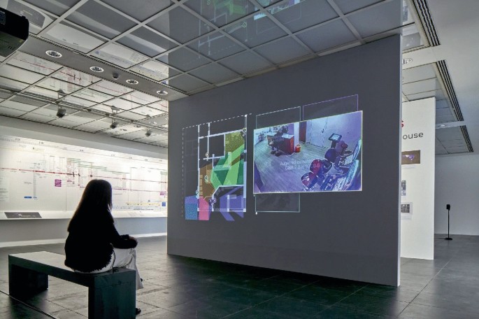 A photo displays a woman sitting and looking at a monitor on the wall, which has a collage or patterns and photos.