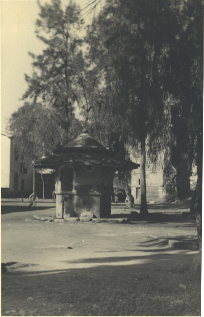 An old photograph of a small hut like structure placed in the center of a town, with taller buildings in the background. A large tree provides shade over the hut like structure, which has a small window on one side.