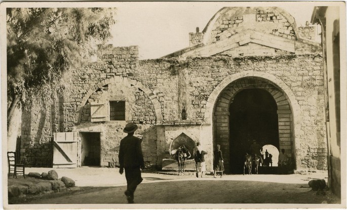 An old photograph of a large brick building, with a large arched entrance. A horse rider and a few people on foot pass under the archway.