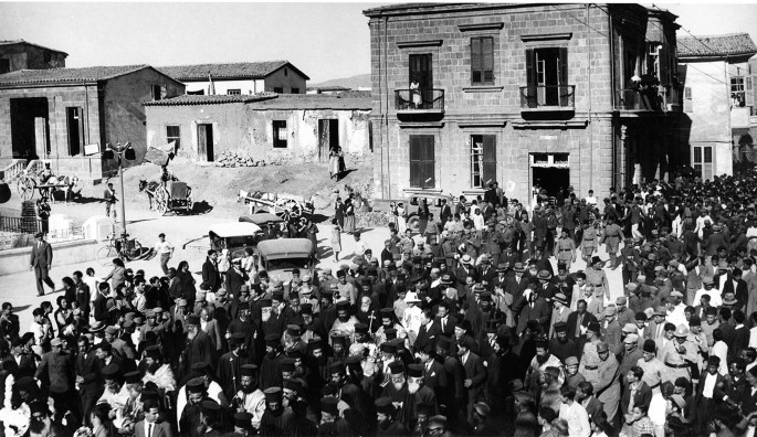 An old photograph of a crowd of people walking in procession down a street. A group of priests is in the center of the procession. Behind the crowd are a few brick buildings, and a few donkey drawn carts.