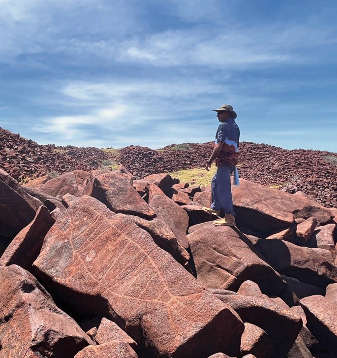 A photo of Peter Cooper standing on a rocky place. A large block in the front has petroglyph of a fish.