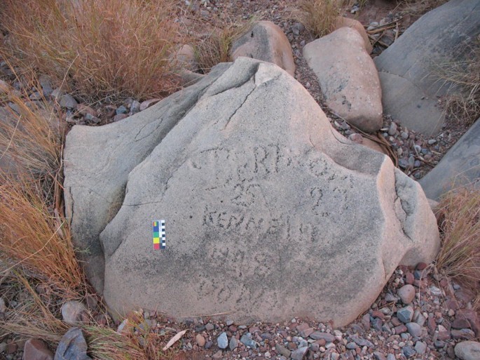 A photograph depicts a rock with inscriptions found in the Roma Gorge boulder, surrounded by dried grass.