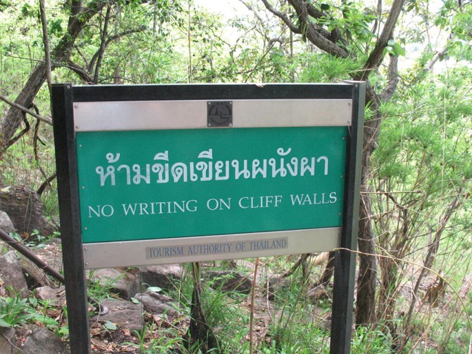 A photograph of a signboard at the Pha Phak Wan rock art site against the backdrop of the jungle. Text on the board reads, no writing on cliff walls, along with a translation in a foreign language.