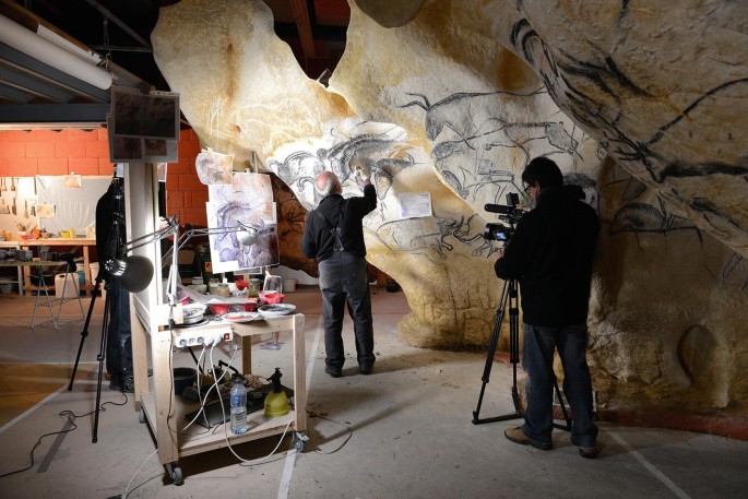 A photograph of an artist painting on a rock formation while another person beside films it.