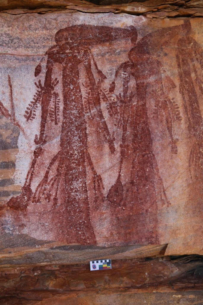 A photograph of a rock face with paintings of a giant creature with shoulder arm and wrist ornamentations and waist band.