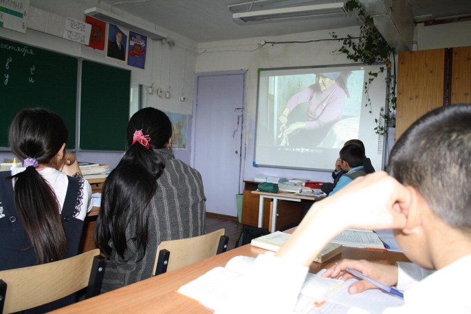A photo of a classroom with students watching a projector screen situated on the side wall of a classroom. A blackboard is positioned in front, surrounded by several charts. Below the screen, there is a table with stacks of books.