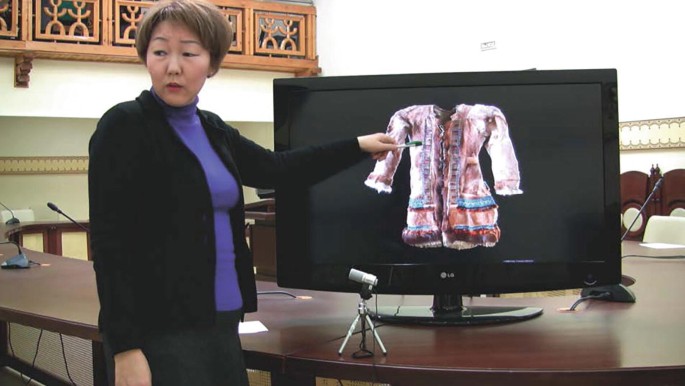 A photo of a woman in a formal suit points to a monitor screen that displays a garment.