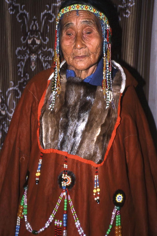 A photo of an old woman dressed in Inuit Parka with a cloth made of fur around her neck. She also wears hairbands around her head with hanging beads.