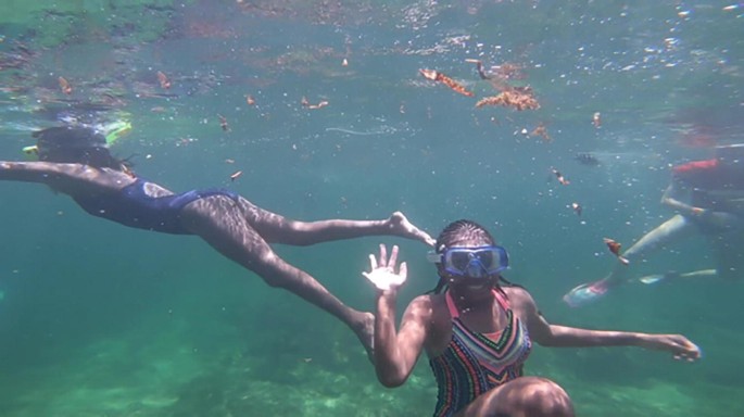 A photograph of a group of people snorkeling underwater, exploring the clear green waters. There are small fish in the background.